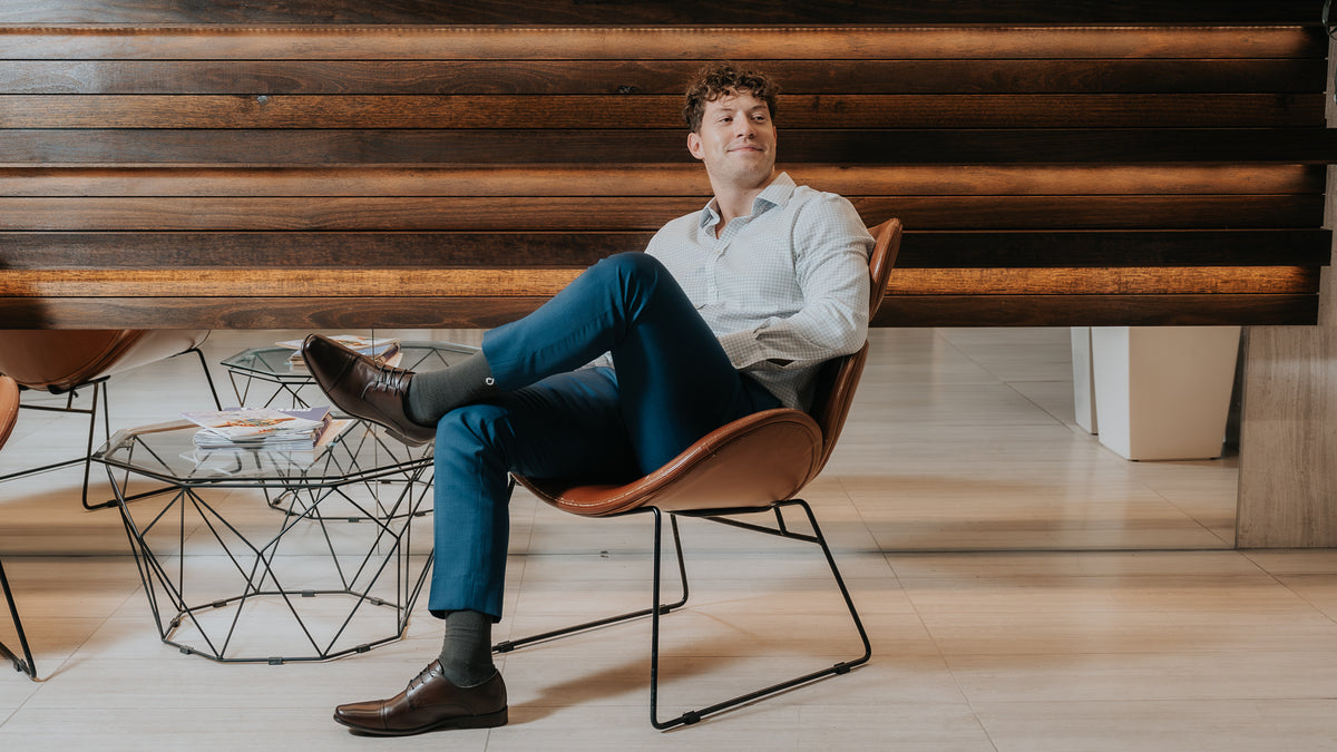 Man in a blue suit and brown dress shoes wearing dark grey DryStep socks, seated in an office lobby