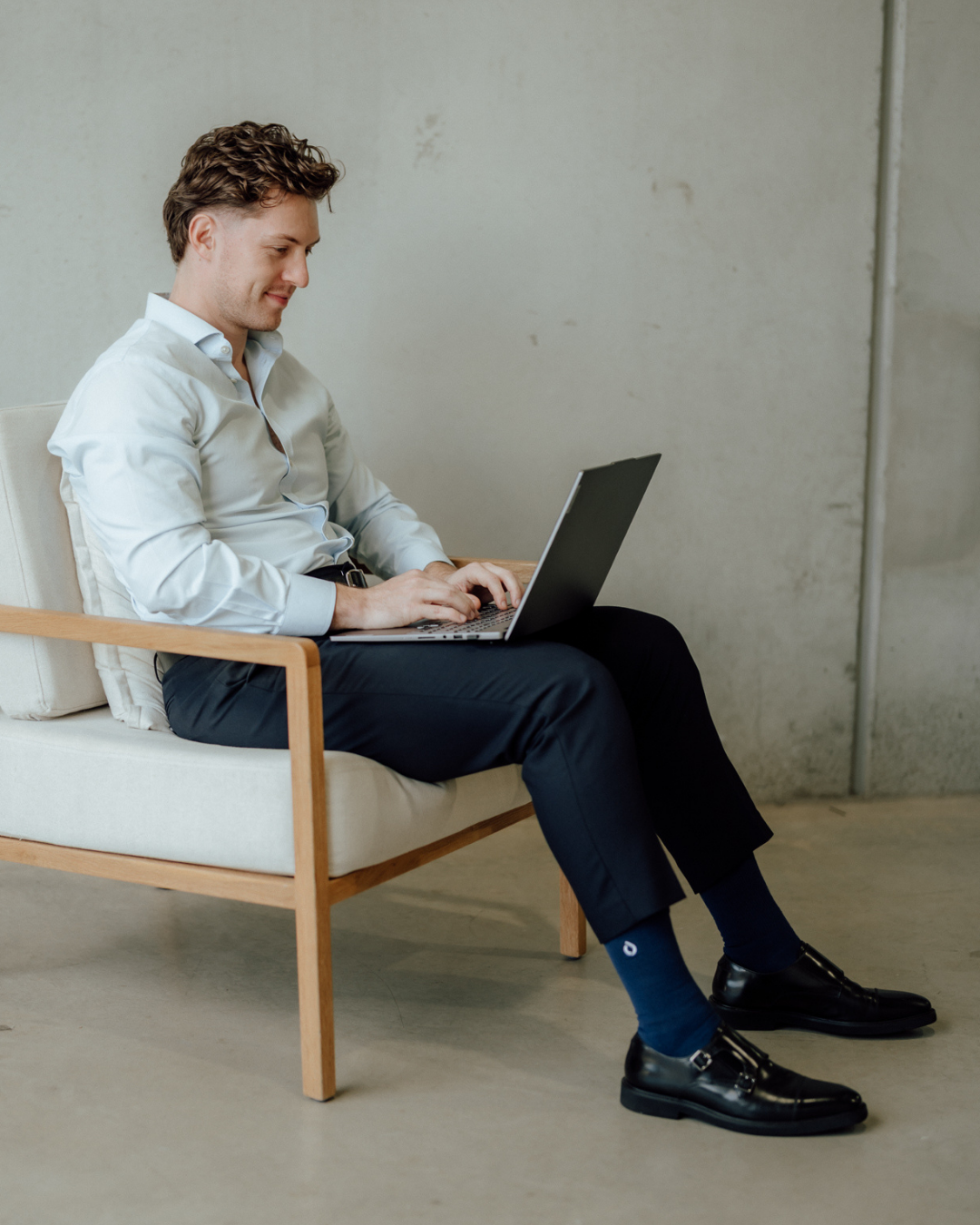 A man sits in a white armchair, typing on a laptop wearing midnight navy drystep socks