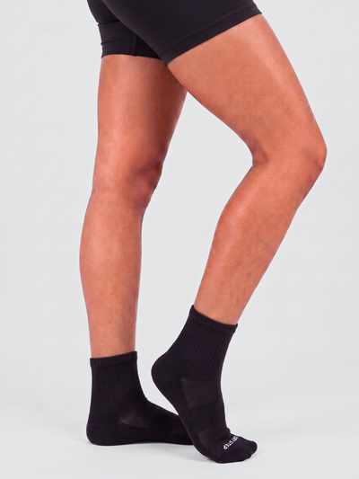 Black athletic quarter socks worn on a female model's feet, shown from the waist down against a light grey studio background.